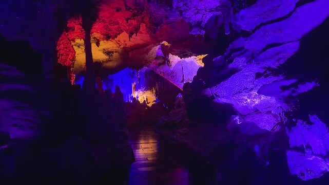 Underground cave system with colorful LED illumination. Stalactites and stalagmites glowing in purple and orange. Natural geological formation for tour exploration.