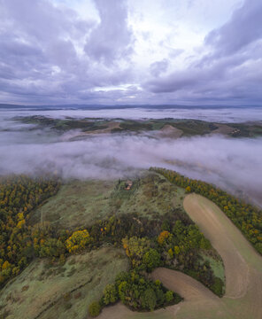 Aerial view of rolling hills and fields in the Tuscan countryside covered in a thick layer of morning mist under a cloudy sky, Tuscany, Italy.