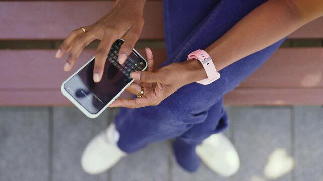 A person sits on a bench and uses their smartphone with fingers on the screen. The location has light paving and the person is wearing a casual outfit.