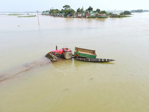 Aerial view of a red tractor stranded on a small patch of land surrounded by floodwater with a man standing on a boat attached to the vehicle in Sunamganj, Sylhet Division, Bangladesh.