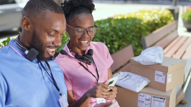 Two workers smile while resting outside a delivery hub. They check their phones and talk to each other while packages are stacked nearby. It is a sunny day.