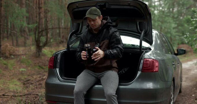 Man sitting on car trunk and drinking coffee from thermos with audio included