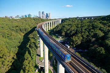 Fototapeta premium A metro train of Taoyuan Airport MRT travels on the elevated rail, crossing green wooded hills on a beautiful sunny day with high rise buildings in Linkou Dist. in backgroud in New Taipei City, Taiwan