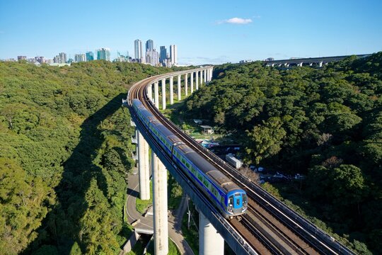 A metro train of Taoyuan Airport MRT travels on the elevated rail, crossing green wooded hills on a beautiful sunny day with high rise buildings in Linkou Dist. in backgroud in New Taipei City, Taiwan