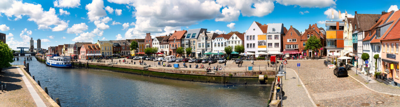 Panoramic view of Husum's historic inner harbor with restaurant ship, waterfront promenade, colorful gabled houses and lively city center.