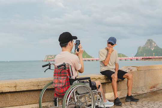 Back view of a young disabled man in a wheelchair taking a photo of his male caregiver posing at the beach with a smartphone, enjoying a joyful and inclusive outdoor travel lifestyle.
