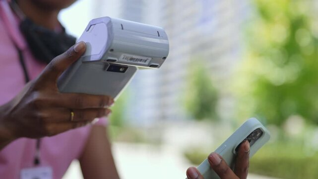 A person is in a park and using a device to scan a phone. The sun is out and there are other people walking and enjoying the area. Trees surround the path where they sit.