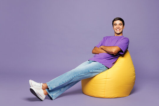 Full body young smiling happy man wear violet t-shirt casual clothes sit in bag chair hold hands crossed folded look camera isolated on plain pastel light purple background studio. Lifestyle concept.