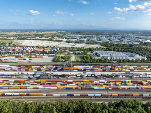 Munich, Germany - 20 July 2026: Aerial view of Container Terminal Munich with rows of colorful shipping containers, freight trains, and industrial infrastructure under a bright blue sky.