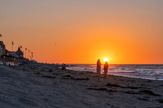 Saint-Aubin-Sur-Mer, France - 07 26 2026: Two women standing in front of the sun on the beach with the jetty and the sea around at golden hour