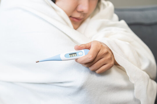 Close-up of sick Asian woman hand holding a digital thermometer showing high body temperature of 39.7 degrees celsius, female wrapping in white blanket having a severe fever at home