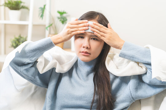 Sick young Asian woman applying cooling gel patch sheet on her forehead suffering from high fever, headache and migraine while resting under blanket on sofa at home
