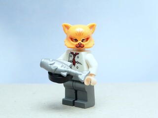 Fototapeta premium Indonesia - May 16, 2025 : Mini toy of action figure at table with blurred background. Miniature photography concept design. Minifigure of orange cat character wearing chef costume with pan and fish.