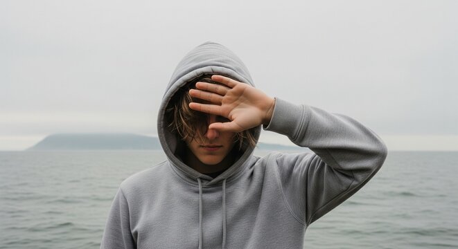Young Person in Grey Hoodie Covering Face with Hand Against Overcast Sky and Ocean Background