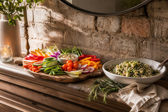 Mantel with board of colorful sliced raw vegetables, quinoa avocado salad, rosemary, linen, rustic brick, clean eating gathering concept