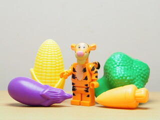 Fototapeta premium Indonesia - February 10, 2026 : Mini toy of action figure at outdoor with blur background. Miniature photography concept design. Minifigure of tiger guy character with eggplant, corn, carrot, broccoli