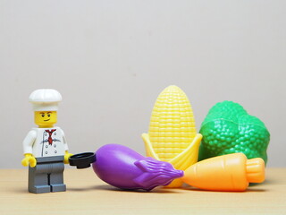 Fototapeta premium Indonesia - February 10, 2026 : Mini toy of action figure at outdoor with blur background. Miniature photography concept design. Minifigure of chef character with eggplant, corn, broccoli and carrot.