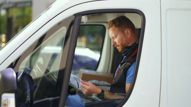 A delivery driver sits in a white van and checks packages. The driver looks at documents and prepares for deliveries. The setting is an urban environment during daytime.