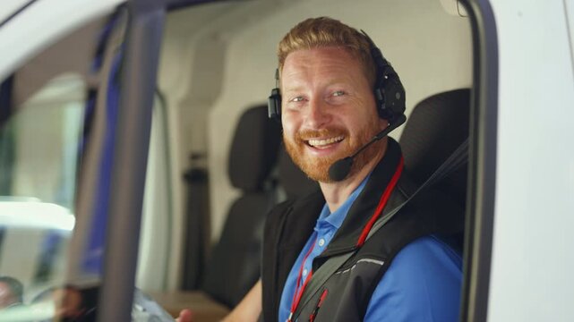 A man sits inside a delivery van, using a headset and reading documents. He is focused on his work while managing tasks. The setting is the interior of the van.