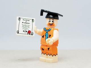 Obraz premium Indonesia - November 24, 2025 : Mini toy of action figure at table with blurred background. Miniature photography concept design. Minifigure of Fred Flintstone with graduation hat and certificate.