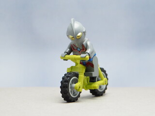 Obraz premium Indonesia - April 14, 2025 : Mini toy of action figure at table with blurred background. Toy photography concept design. Minifigure of Ultra Man character from japanese fiction film riding bicycle.