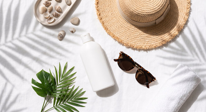 Summer beach flat lay with straw hat sunglasses sunscreen bottle and palm leaves on white towel.