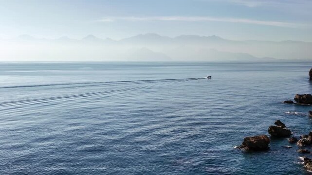 Small boat leaves a trailing wake across calm blue sea near rocky cliffs in antalya.