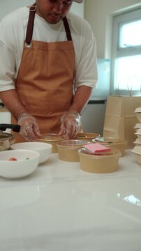 Close-up of a male chef packing freshly prepared fresh meals for online food delivery in a small business kitchen. Cloud kitchen, local restaurant, food startup. Meal prep and entrepreneur concept.
