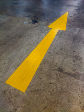 Yellow directional arrow painted on a basement parking floor, guiding drivers along designated routes and improving vehicle navigation, circulation, and safety within the parking facility.