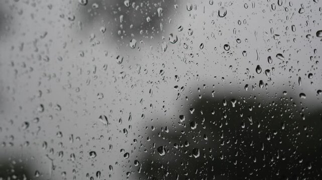 Close up macro view of rain drops sliding and trickling down a glass window pane on a rainy day, with a soft blurred background creating a calm and moody atmosphere.