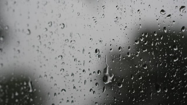 Close up macro view of rain drops sliding and trickling down a glass window pane on a rainy day, with a soft blurred background creating a calm and moody atmosphere.