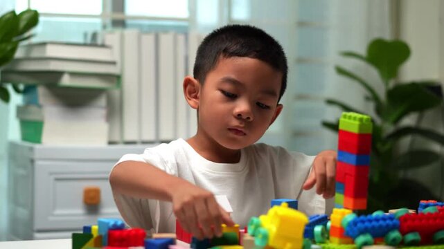 Child builds colorful structures with toy bricks. Focused expression shows quiet concentration. Soft indoor lighting adds warm, playful atmosphere.