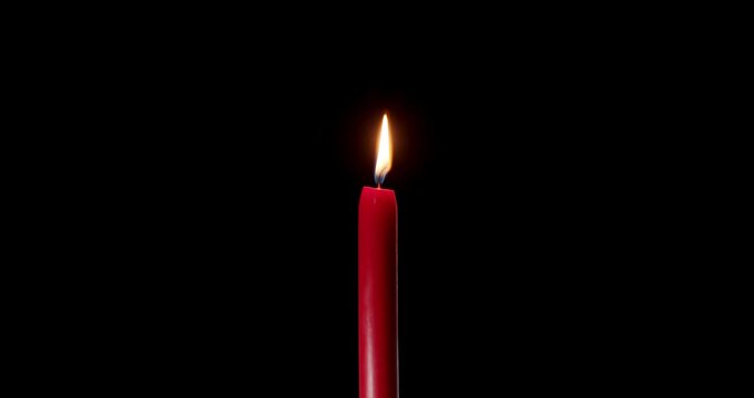 Brightly lit red candle burning with a warm glow in the dark. A striking wax candle that contrasts with a completely black background. This classic candle represents memory and celebration.