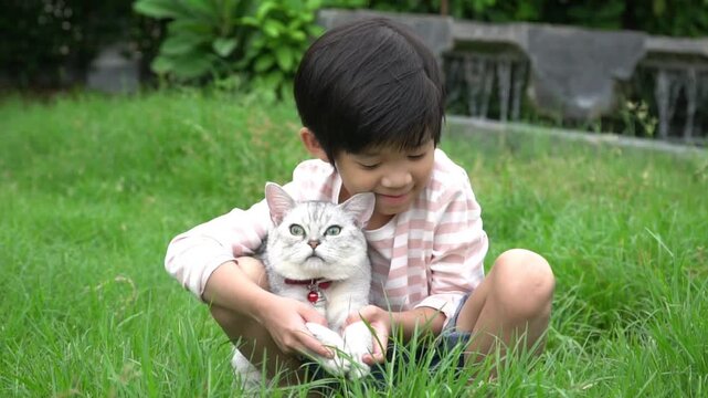 Cute Asian boy hugging and playing with a silver cat on green grass outdoors.