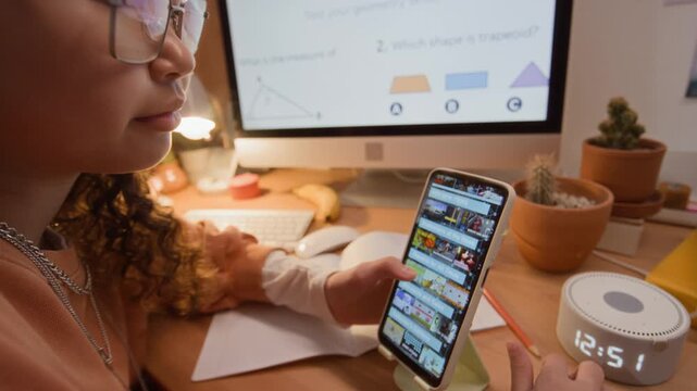 Asian teenager wearing headphones and glasses scrolling on smartphone while studying geometry at desk. Footage suitable for promoting e-learning apps and online education platforms