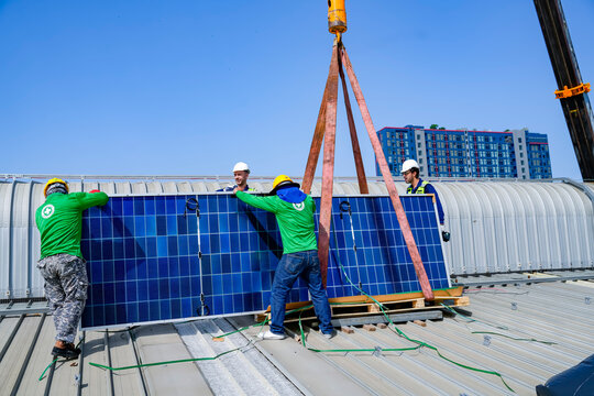 Workers install solar panels on a building roof in bright sunlight using a crane in an urban area