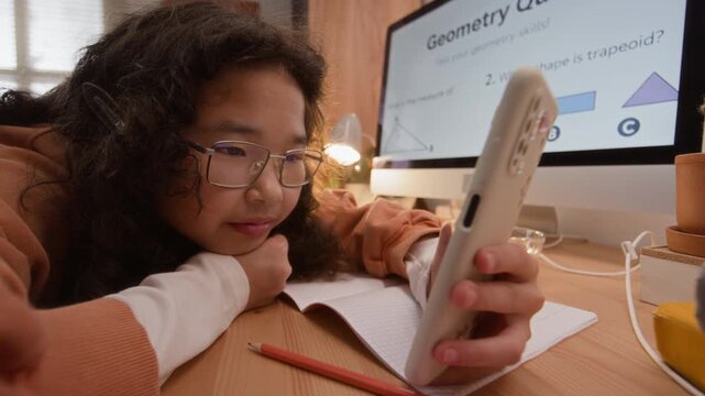 Asian teenager lying on desk, scrolling on smartphone instead of studying geometry on computer screen. Footage represents online learning challenges and digital distraction in education