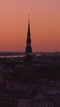 Aerial dusk pan centers St. Peter's Church spire with rooster vane in Riga. The Daugava River, cranes, and old town rooftops appear as tower windows glow.