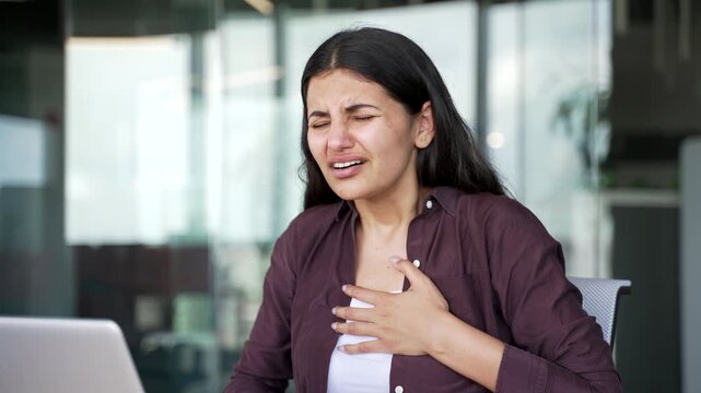 Young businesswoman suffering from intensive chest pain while working on laptop in modern office. Female employee holds her chest, experiencing discomfort, breathing difficulties, heart attack. Close 
