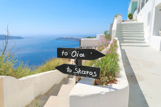 Black wooden signs point toward Oia and Skaros along a whitewashed walkway overlooking the Aegean Sea in Santorini, Greece &mdash; a classic Cycladic travel scene.