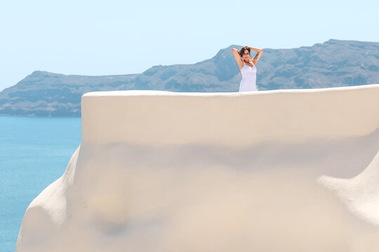A woman stands on a sunlit terrace framed by volcanic cliffs and blue sea in Santorini, Greece &mdash; a tranquil moment of summer grace.