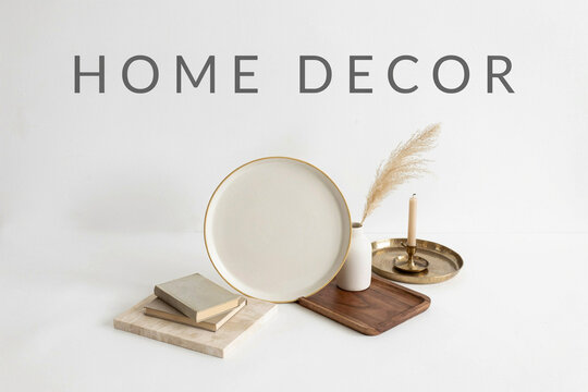 An elegant arrangement of various home decor items including a large cream plate a small white vase with pampas grass a lit candle and a stack of books creating a stylish interior display