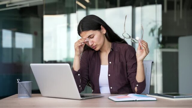 Tired businesswoman experiences eye strain, fatigue or headache after long hours of computer work in modern office. Female employee removes glasses rubs her eyes while taking break from laptop screen.