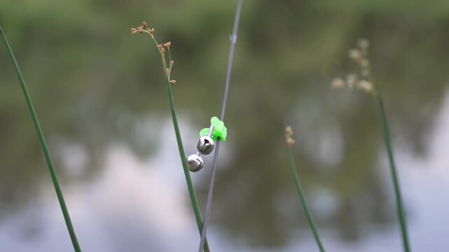Close-up 4K footage of a small fishing bite alarm bell attached to the tip of a fishing rod beside a pond. The bell is used as a simple bite indicator to alert an angler when a fish pulls the line.
