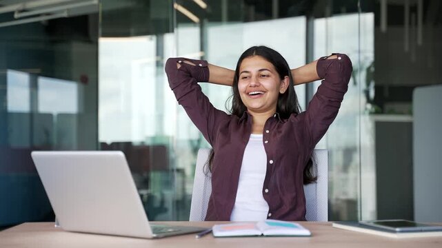 Happy young businesswoman finishes work on laptop while sitting at desk in modern business office. Relaxed female manager leans back in chair, feeling satisfied after completing daily tasks, projects.