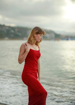 Elegant young woman in red dress, holding glass of wine, having fun and enjoying her vacation on seashore with the city in background