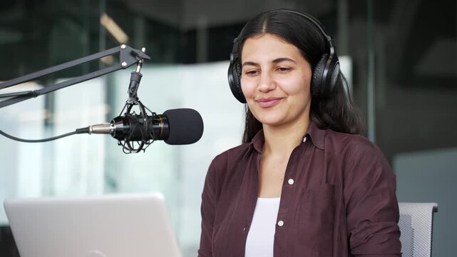 Young businesswoman hosting online podcast speaking into microphone with laptop sitting in office. Professional female content creator records business discussion, communicates with audience. Close up
