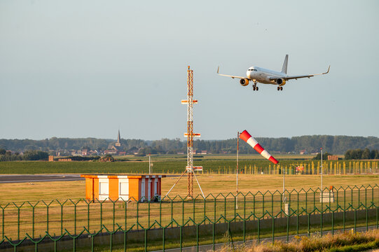 A passenger aircraft descends on final approach beyond the approach lighting and windsock at Brussels Airport in Belgium in warm evening light.