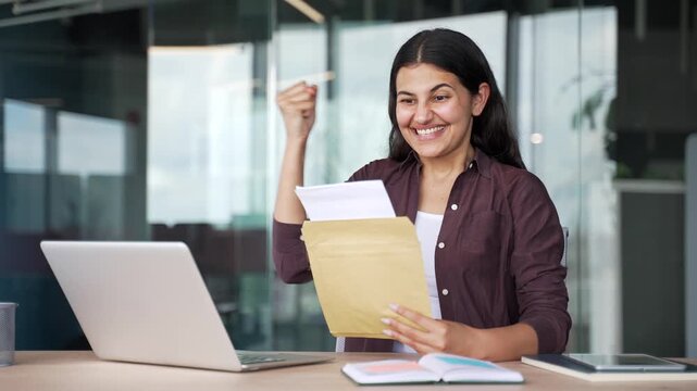 Happy young businesswoman receives good news approval letter while sitting in modern office. Smiling female employee is satisfied with positive notification, celebrates success, exciting achievement.