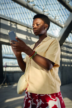 Vertical shot of young Black woman checking smartphone while commuting, managing mobile communication and digital tasks. Ideal for app marketing, connectivity, urban lifestyle